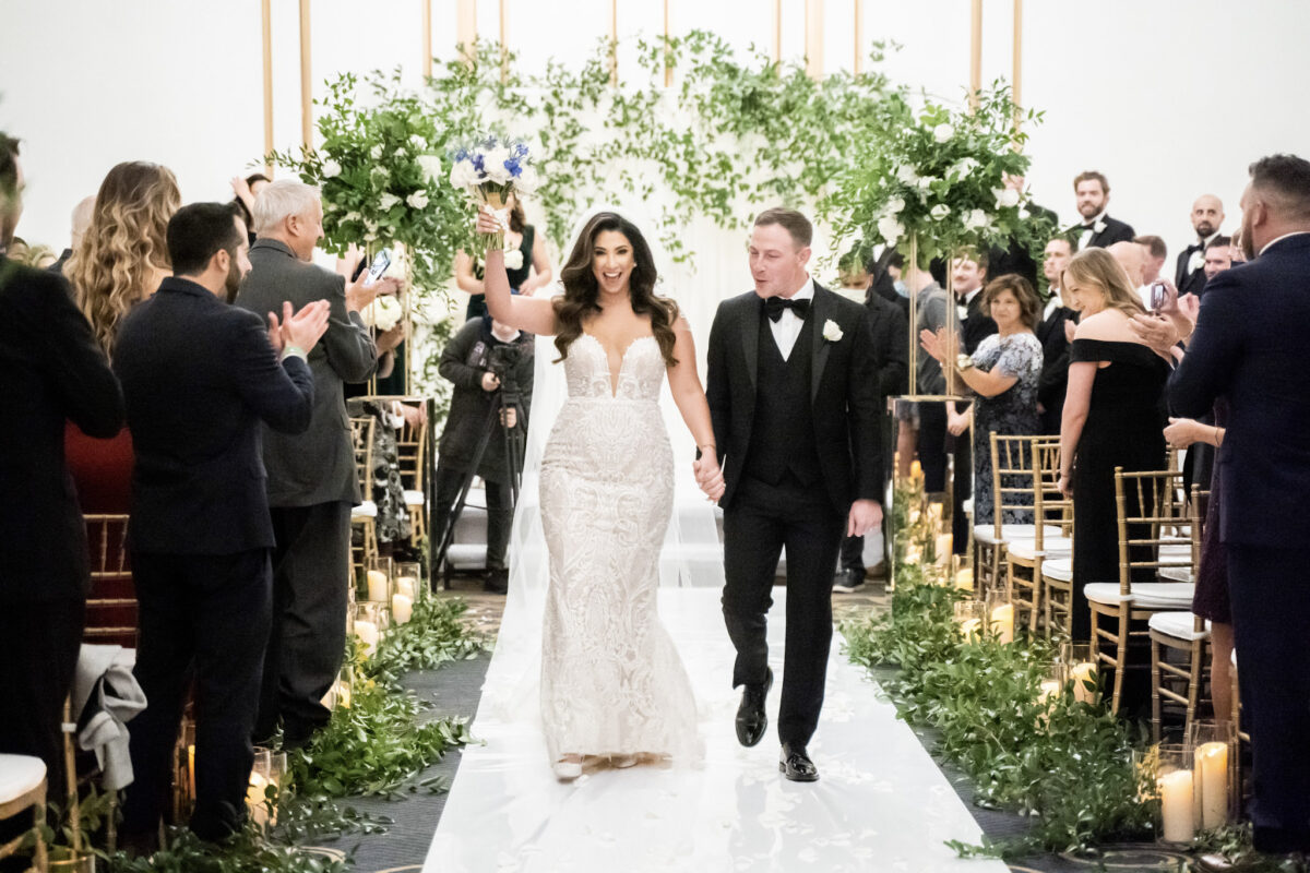 Bride and Groom at indoor wedding ceremony with abundant greenery down the aisle and on the ceremony arch.