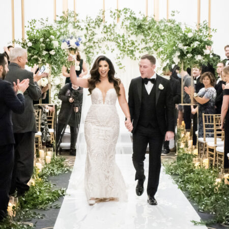 Bride and Groom at indoor wedding ceremony with abundant greenery down the aisle and on the ceremony arch.